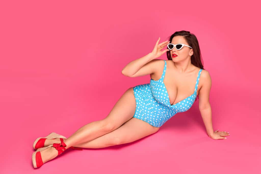 A woman in a blue polka dot bikini lays on a pink background, showcasing size-inclusive fashion.